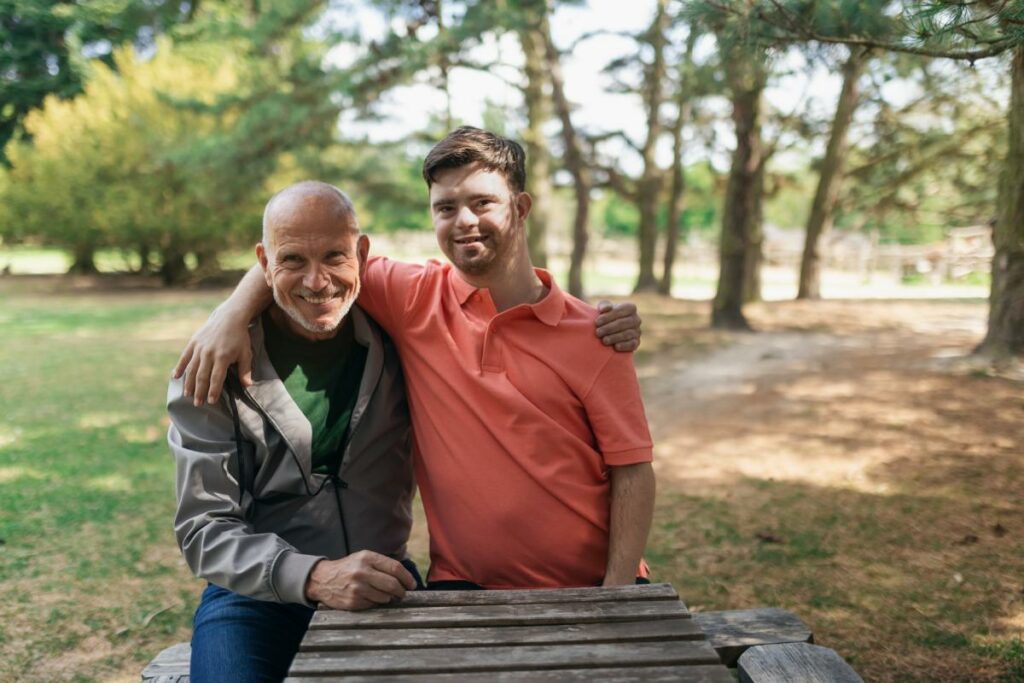 An old man wearing a light jacket and a younger adult male wearing an orange golf shirt with an arm around each other's shoulder in the woods