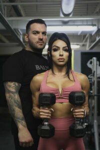 James Ayotte standing behind a woman he is training holding two dumbbells in pink workout clothing
