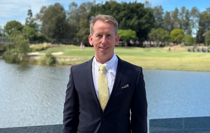 John Margerison in a suit standing in front of water with a park behind it