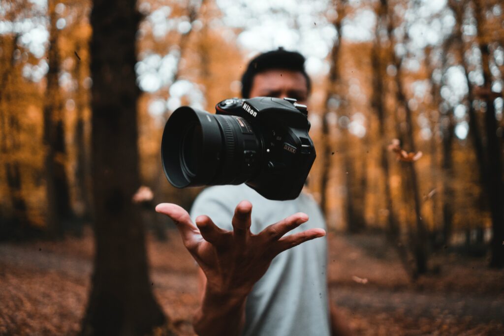 A man in a blue shirt tossing a camera in front of his face in the woods