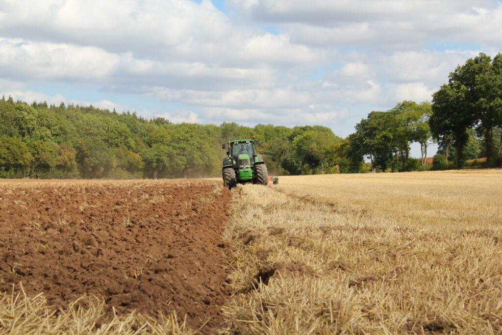 A tractor plowing a farmer's field with a forest and a cloudy blue sky in the background