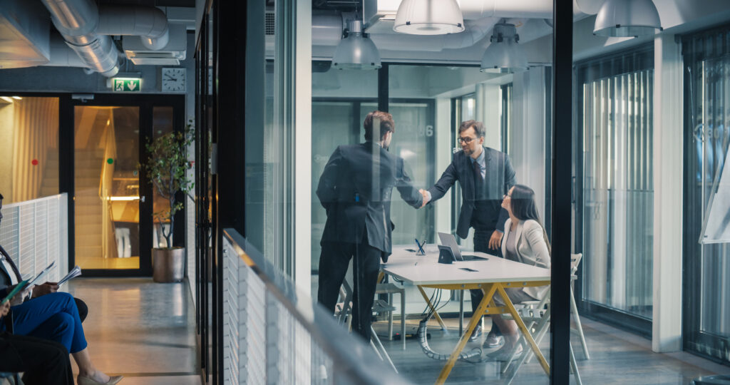 Business Leaders Complete a Job Interview With a Confident Handshake in a Glass Meeting Room, While a Hiring Manager Reviews Notes and Additional Candidates Wait in the Hallway