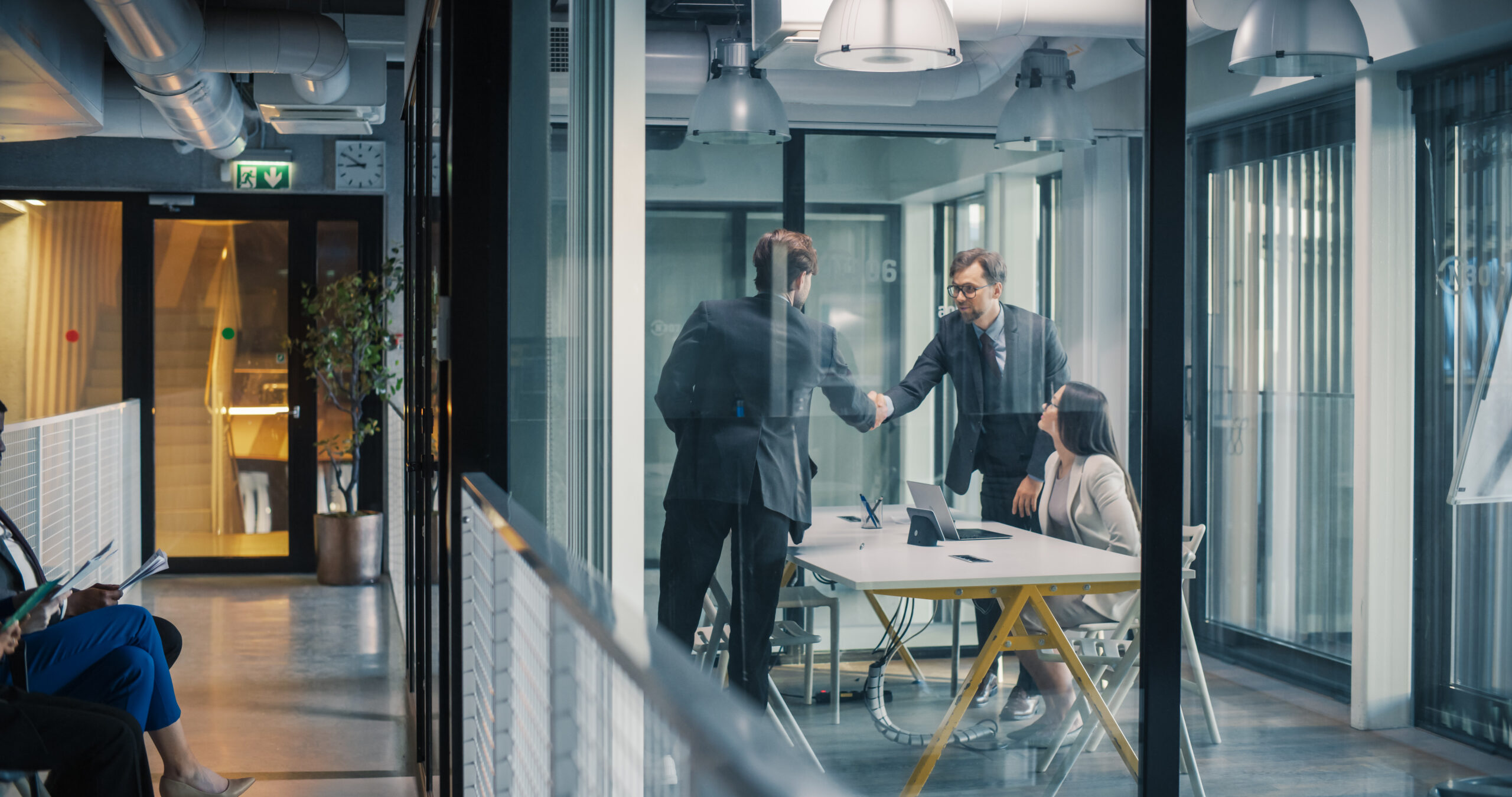 Business Leaders Complete a Job Interview With a Confident Handshake in a Glass Meeting Room, While a Hiring Manager Reviews Notes and Additional Candidates Wait in the Hallway