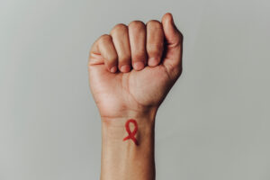 the raised fist of a man with a red awareness ribbon for the fight against AIDS painted in his wrist, against an off-white background with some blank space around him